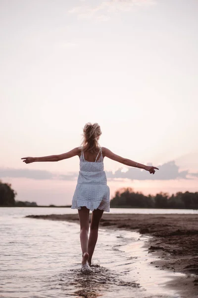 Hermosa mujer en la playa vista trasera. Una joven rubia esbelta con un vestido blanco está girando y bailando en la orilla del río sobre el fondo de la puesta del sol y con las manos levantadas —  Fotos de Stock