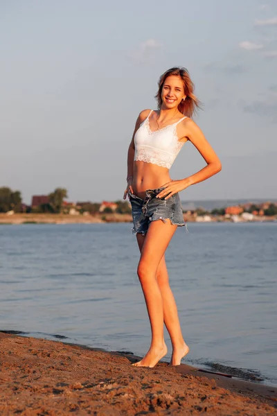 Женщина на пляже. Красивая улыбающаяся молодая блондинка в кружевном белом топе и расстегнутых джинсовых шортах стоит и позирует на песке у воды — стоковое фото