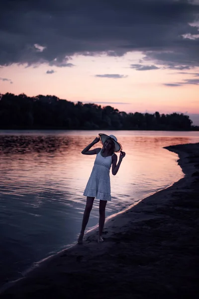 Eine junge Frau vor dem Hintergrund eines Flusses und Wolken. Eine schlanke, schöne Blondine steht in einem weißen Kleid am Ufer des Flusses und hält einen weißen Strohhut in den Händen. — Stockfoto