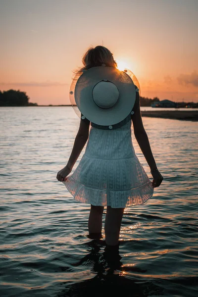 年轻女子背景在夕阳的后视镜.照片上美丽的金发姑娘穿着白衣，脖子上戴着一顶草帽，站在水里，双手托着裙子的边沿 — 图库照片