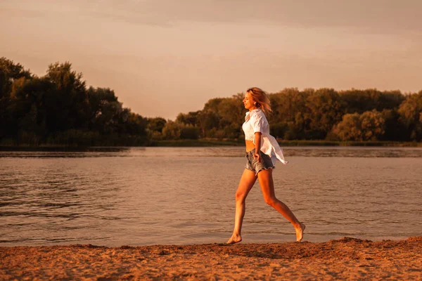 Młoda kobieta biegnąca nad rzeką o zachodzie słońca. Profil zdjęcie pięknej szczęśliwej blondynki w białych ubraniach i jeansowych spodenkach biegających po piasku w malowniczym krajobrazie — Zdjęcie stockowe