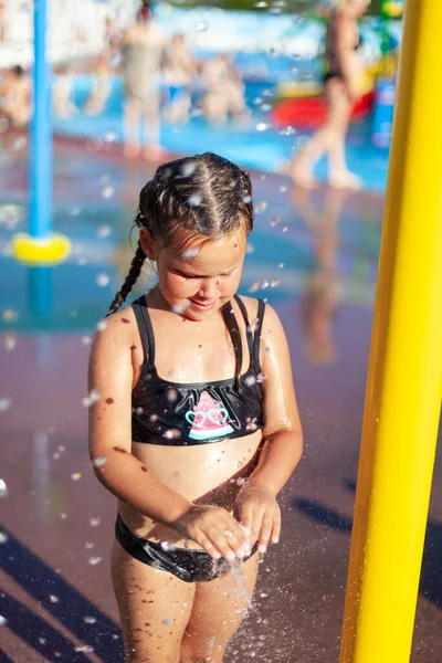 Malé dítě si hraje u bazénu. Vyčiněná dívka s copánky stojí u fontány parku a mokré ruce ve vodě na pozadí aquaparku v horkém letním dni — Stock fotografie