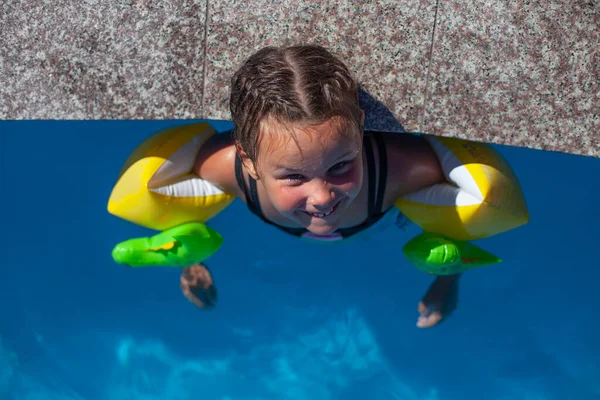A gyerek arcokat vág a medence szélén. Boldog kislány felfújható karszalagban mosolyog és élvezi a hétvégét a vízi parkban, felülnézet — Stock Fotó