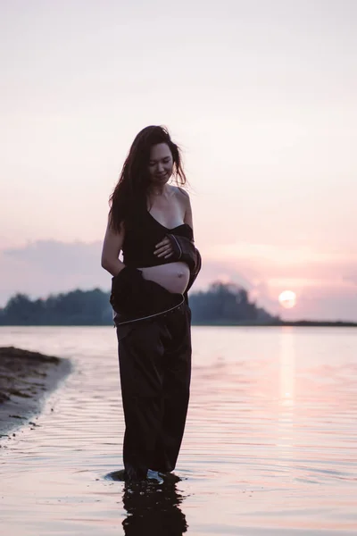 Mujer embarazada sonríe. Hermosa joven embarazada en traje oscuro y pelo largo y castaño se para en la orilla del mar en rayos de sol poniente y sostiene su estómago con las manos —  Fotos de Stock