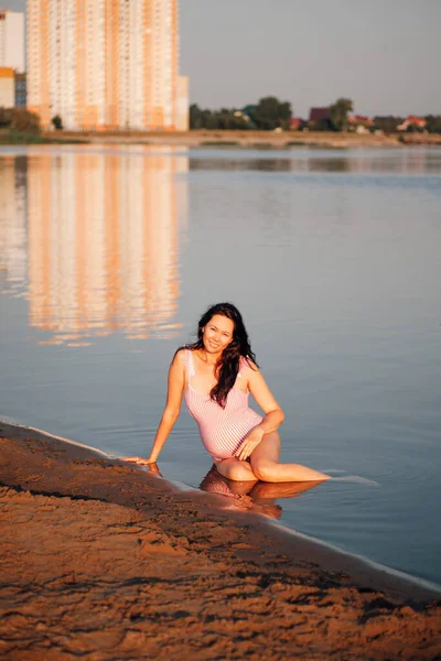 Беременная женщина на пляже, милая, расслабляющая молодая беременная женщина в розовом купальнике наслаждается прохладой речной воды в жаркий летний день на закате — стоковое фото