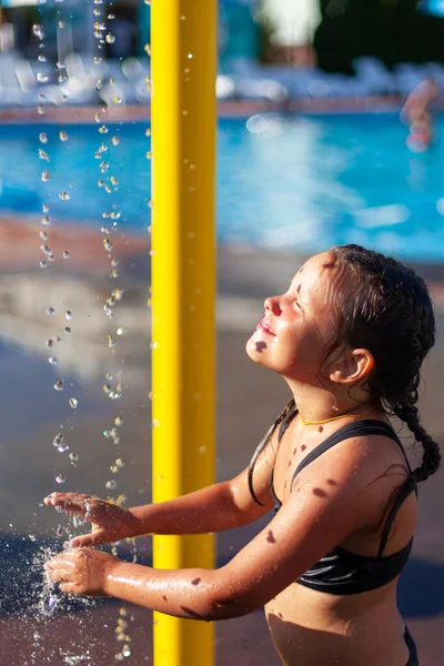 Kind spielt im Wasserfall. Kleines glückliches Mädchen in den Strahlen der heißen Sommersonne steht unter Wasserstrahlen in der Nähe des Pools und benetzt ihre Hände im Wasser — Stockfoto