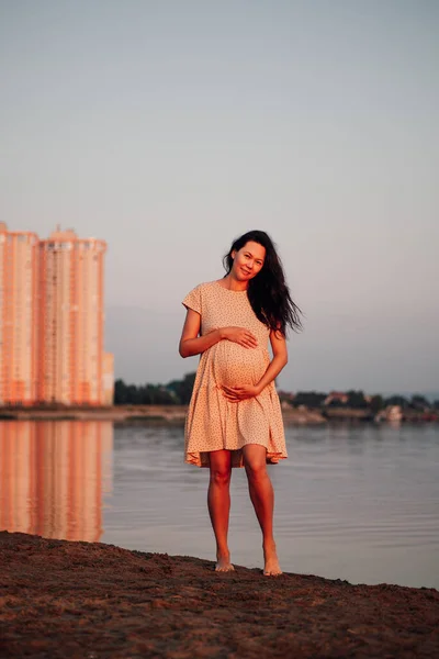 O femeie frumoasă pe malul râului. Femeie asiatică însărcinată cu părul ei care curge în vânt și rochie bej stă pe fundalul clădirilor înalte și își ține stomacul cu mâinile — Fotografie, imagine de stoc