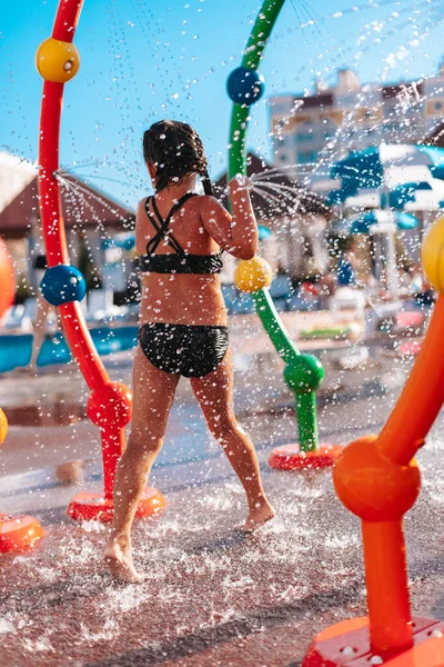 Kleines Mädchen steht mit dem Rücken zum Pool. Glückliches schönes Mädchen im schwarzen Badeanzug spielt während ihres Urlaubs mit Wasserspritzern am Pool des Wasserparks — Stockfoto