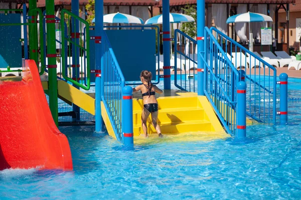 Día soleado de verano en el parque acuático. Niña disfruta de las vacaciones y la recreación en la piscina del centro de entretenimiento, sube los pasos de color amarillo brillante del tobogán acuático —  Fotos de Stock