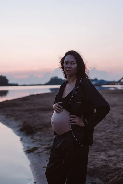 Retrato de una mujer embarazada en la playa. Foto romántica de la atractiva joven morena en pijama oscuro desabotonado en la playa mirando a la cámara y su cabello soplando en el viento —  Fotos de Stock