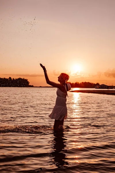 Foto de perfil de mulher salpicando água. Silhueta de bela loira delgada feliz em um vestido de verão e um chapéu de palha com uma fita no fundo do pôr do sol e da praia com as mãos levantadas — Fotografia de Stock