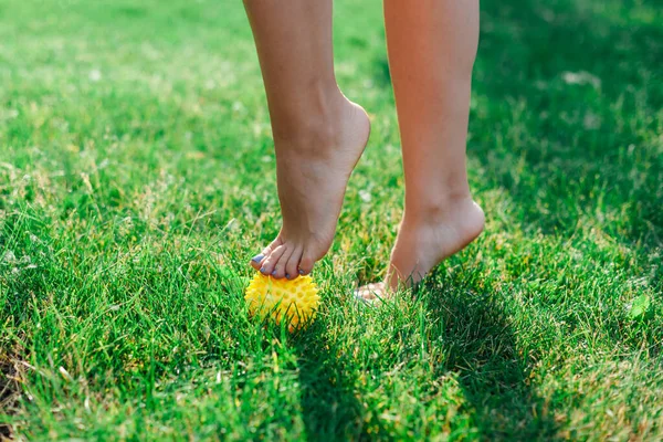 Dettagli dei piedi nudi di una giovane donna in piedi in punta di piedi su una palla gialla di gomma appuntita per rilassare i muscoli e ridurre il dolore su un prato verde in una mattina di sole — Foto Stock