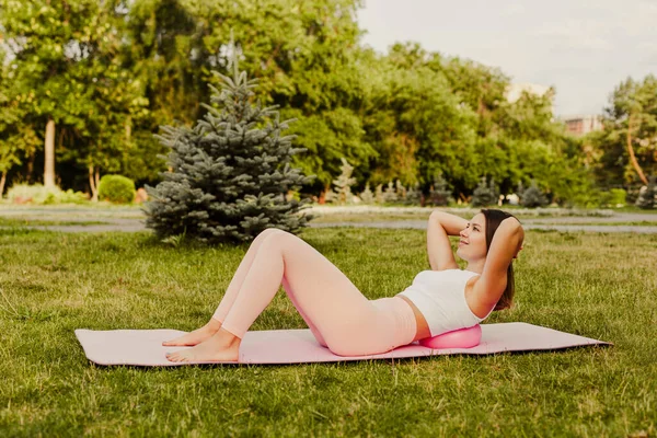 Atlética, mujer esbelta en ropa rosa y pequeña bola de goma haciendo Pilates en estera rosa al amanecer en el parque en verano — Foto de Stock