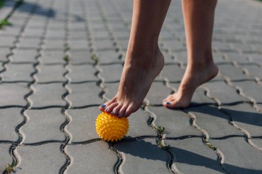 Çıplak ayakla dikenli sarı topa basan genç bir kadının detayları kasları gevşetmek ve güneşli bir günde döşeme taşındaki acıyı azaltmak için.