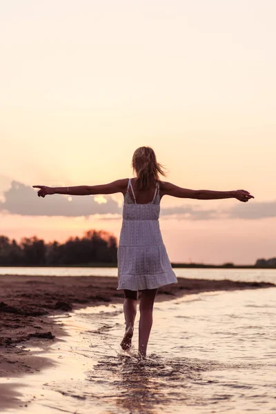 美丽的女人在海滩后视镜。一位身穿白衣、身材苗条的年轻金发姑娘，在夕阳西下的背景下，双手高举，沿着河岸奔跑 — 图库照片