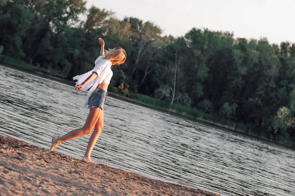 Młoda kobieta tańczy na plaży. Szczęśliwa piękna blondynka w białej koszuli i top i jeansowe szorty na tle krajobrazu nad rzeką — Zdjęcie stockowe