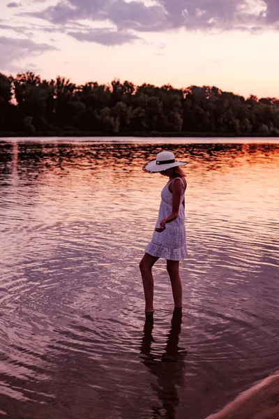 Eine Frau mit Strohhut bei Sonnenuntergang. Eine schöne junge schlanke Blondine steht im weißen Sommerkleid im Wasser und hebt die Arme zur Seite — Stockfoto