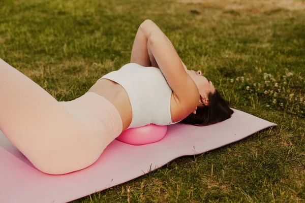 close-up of slender woman in pink top hugging arms around shoulders and lying with shoulder blades on small rubber ball in park in summer, concept of body care and slow life