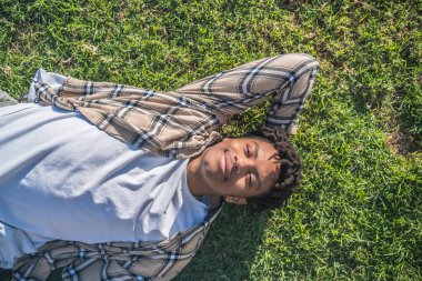 Siyah afro amerikan adam elinde tabletle parktaki bej ekose ceketli yeşil çimlerin üzerinde oturuyor eski kasabadaki sıcak renkler