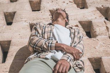 Afrikalı Amerikalı siyahi bir adam, eski kasabadaki bir parkta bej renkli ekose ceketli bir duvarın önünde poz veriyor.