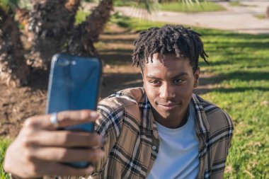 Siyah afro amerikan adam elinde tabletle parktaki bej ekose ceketli yeşil çimlerin üzerinde oturuyor eski kasabadaki sıcak renkler