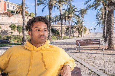 Afro-Amerikalı, sarı kazaklı siyah adam parktaki ahşap bankta uzanan akıllı telefona bakıyor.