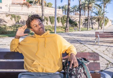 Afro-Amerikalı, sarı kazaklı siyah adam parktaki ahşap bankta uzanan akıllı telefona bakıyor.