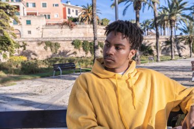 Afro-Amerikalı, sarı kazaklı siyah adam parktaki ahşap bankta uzanan akıllı telefona bakıyor.