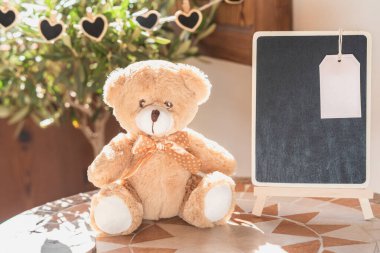 Sevgililer günü oyuncak ayısı yanında karatahtayla mozaik masasında ve arka plandaki pencereye kalpler yerleştiriyor.