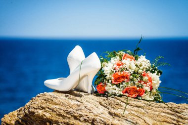 Beyaz düğün ayakkabıları ve gelin buketi beyaz, yeşil ve kırmızı renklerle kayaların üzerinde mavi gökyüzü ve bulanık arka planda deniz var.