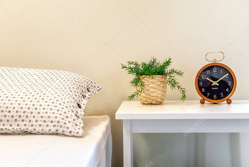 Bedside table with greenplant in straw pot and clock on it. Part of bed is visible. Interior details. light colours. 