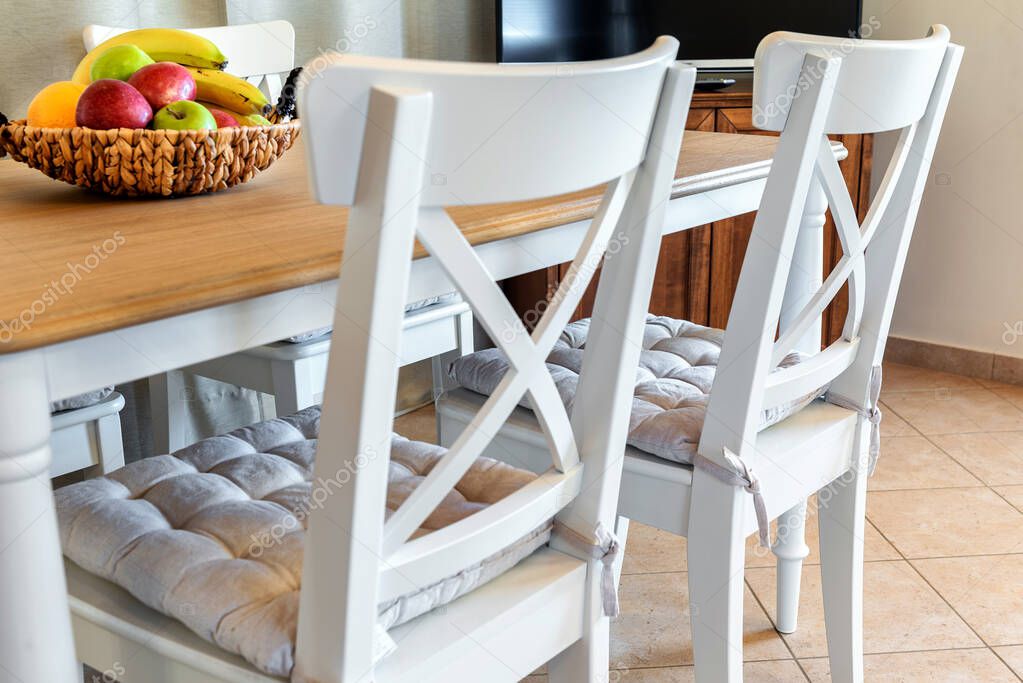 Part of dining room. Two white chairs in selective focus and part of the table with some fruits on it. 