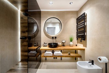 Bej ve kahverengi sıcak renklerde modern fayanslı banyo. Cam bölmeli duş kabini, siyah lavabo için ahşap standı ve duvarında yuvarlak ayna var. Seçici odak