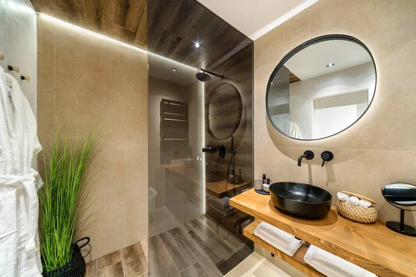 用米黄色和棕色暖色调装饰的现代瓷砖浴室 它有带有玻璃隔板的淋浴房 黑色水池的木制支架和墙上的圆形镜子 — 图库照片