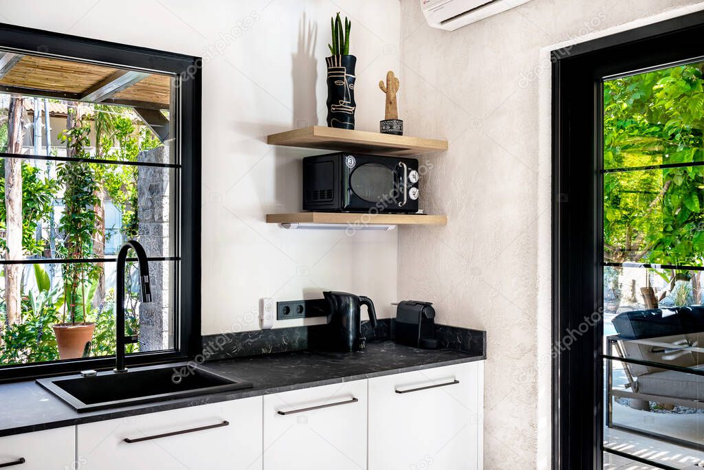 Modern kitchen interior in the  apartment villa. Black marble, quartz counter top kitchen with black sink and faucet, basic electrical equipmen, window overlooking to patio garden. Selective focus.