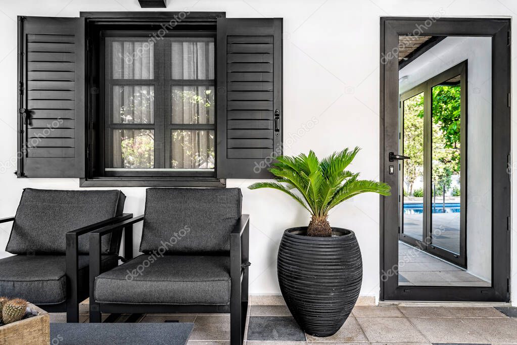 Outdoor terrace. Dark grey color armchairs, flower pot and window shutters with the white wall at the background. 