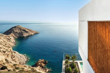 Sakin turkuaz deniz ve kayalık uçurumlara bakan modern kıyı mimarisi. Minimum beyaz bina ahşap detaylar ve teras bahçesi ile Akdeniz manzarası harmanlanmış..