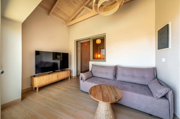 Светлая гостиная с мягким серым диваном, деревянным журнальным столиком, телевизором и лампой из ротанга. Современный натуральный интерьер с легкими текстурами дерева и уютной атмосферой.