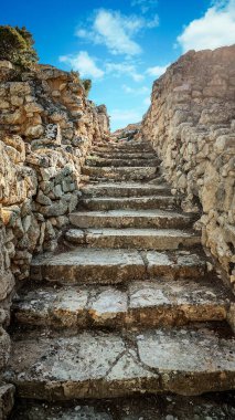 Parlak gökyüzünün altında yıpranmış kaya duvarları arasında yükselen eski taş merdiven, dokulu taşlar, sıcak ışık, engebeli yüzeyler ve yukarıya doğru giden yolu gösteren tarihi bir atmosfer yaratıyor. Eski taş basamaklar