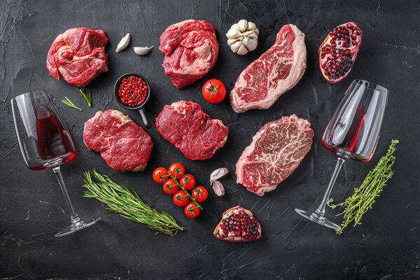 Set of various beef steak cuts, chuck roll, rump andtop blade steak with herbs, seasoning and red wine glass on black table, top view