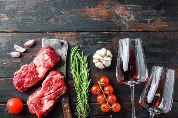 Organic raw chuck eye roll beef steak over old butcher cleaver near red wine glasses with herbs and seasonings on old dark wooden table. Top view, with space for text