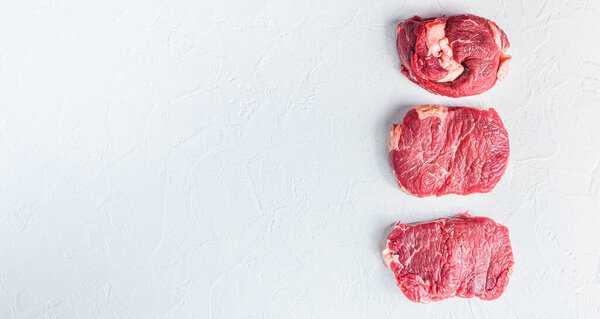 A set of different types of raw beef steaks:top blade, rump, chuck eye roll over white concrete background top view big size banner space for text