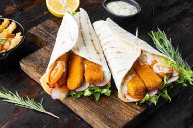 Balık kroketli, peynirli ve sebzeli sandviç, ahşap kesme tahtasının üzerinde, eski, siyah ahşap masa arkasında.