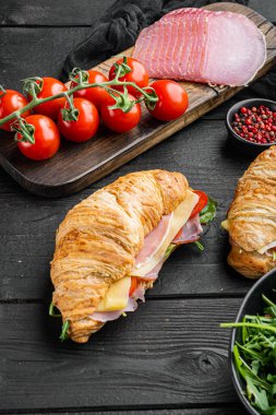 Taze kruvasan ya da salatalı sandviç, jambon, jambon, prosciutto seti, otlar ve malzemelerle birlikte, siyah ahşap masa arkasında.