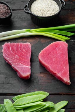 Çiğ ton balığı bifteği, malzemeli taze ton balığı filetosu, yeşil bezelye, susam ve baharat seti, eski siyah ahşap masa arkasında.