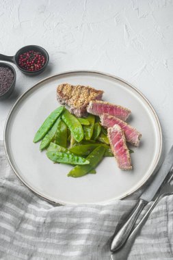 Ton balıklı ızgara biftek, taze soğan ve bezelye, tabakta, beyaz taş zemin üzerinde.
