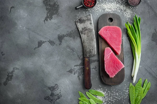 Ahşap kesim tahtasına yeşil bezelye, susam ve otlar dizilmiş çiğ ton balığı parçaları ve eski kasap satır bıçağı, gri taş arka planda, üst manzara düz, metin için yer ve mekan