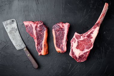 Black Angus Prime çiğ biftek seti, tomahawk, t kemik veya biftek ve sopa biftek, eski kasap satır bıçağı ile, siyah taş arka planda, üst düz yatıyordu