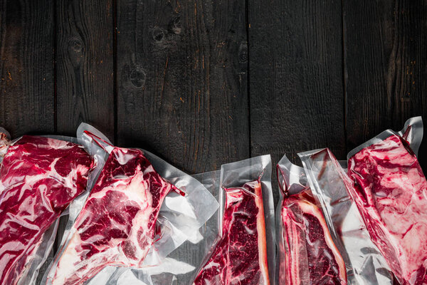 Raw beef meat in the vacuumed skin pack set, tomahawk, t bone, club steak, rib eye and tenderloin cuts, on black wooden table background, top view flat lay, with copy space for text