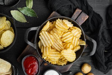 Peynir ve soğanlı farklı patates cipsi çeşitleri, siyah ahşap arka planda, üst manzara düz.